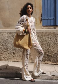 Completo all'uncinetto bianco con motivi floreali, pantaloni dalla vestibilità morbida e una camicia abbottonata coordinata; abbinato a una borsa slouchy color beige.