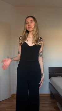 Νεαρή γυναίκα με μακριά ξανθά μαλλιά, φορώντας ένα μαύρο αμάνικο ολόσωμο, στέκεται σε ένα υπνοδωμάτιο με τατουάζ στα χέρια της.