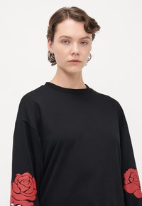 Ung person med kort krøllete hår har på seg en svart langermet skjorte med store røde rose-design på ermene mot en hvit bakgrunn.