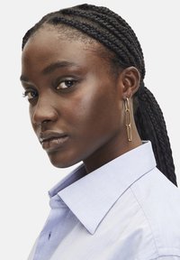 Goldene Kettenohrringe, strukturierte Haut, geflochtenes Haar, trägt ein hellblaues Hemd mit Kragen, vor einem einfarbigen weißen Hintergrund.