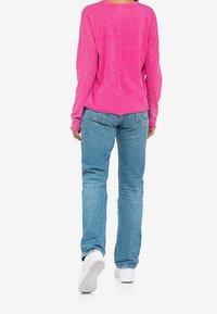 Roze langemouwen top met een relaxte pasvorm, gecombineerd met blauwe spijkerbroeken en witte sneakers. De top heeft een afgeronde zoom en een zachte textuur.