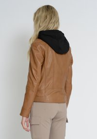 Veste en cuir marron avec une capuche noire attachée, finition texturée, coupe ajustée et détails de couture visibles. Portée avec un pantalon beige.