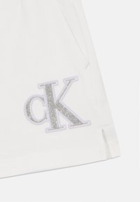 Fehér pamut sort, ezüst csillámos "CK" logóval és kontrasztos lila kerettel, oldalsó hasítékokkal a szabad mozgás érdekében.