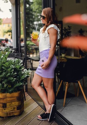 Woman in a white ruffled top and lavender pencil skirt holds a yellow drink. She wears sunglasses and black sandals with a cork sole.
