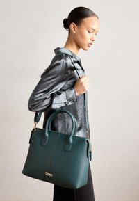 Sac à main en cuir vert avec double poignée et une bandoulière amovible. Comprend des ferrures dorées et une surface texturée.