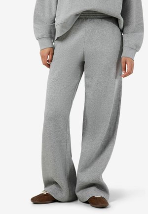 Persona con pantalones deportivos grises sueltos, sudadera gris a juego y zapatos marrones sin cordones, de pie frente a un fondo liso.