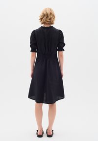 Schwarzes Leinenkleid mit kurzen Puffärmeln und geraffter Taille. Knielang, lockere Passform und weiche Textur. Kombiniert mit schwarzen flachen Sandalen.