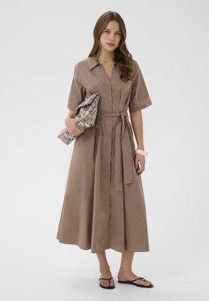 Mujer con un vestido midi marrón con cinturón en la cintura, sosteniendo un bolso de piel de serpiente, llevando una pulsera beige y sandalias negras tipo thong, de pie sobre un fondo blanco.