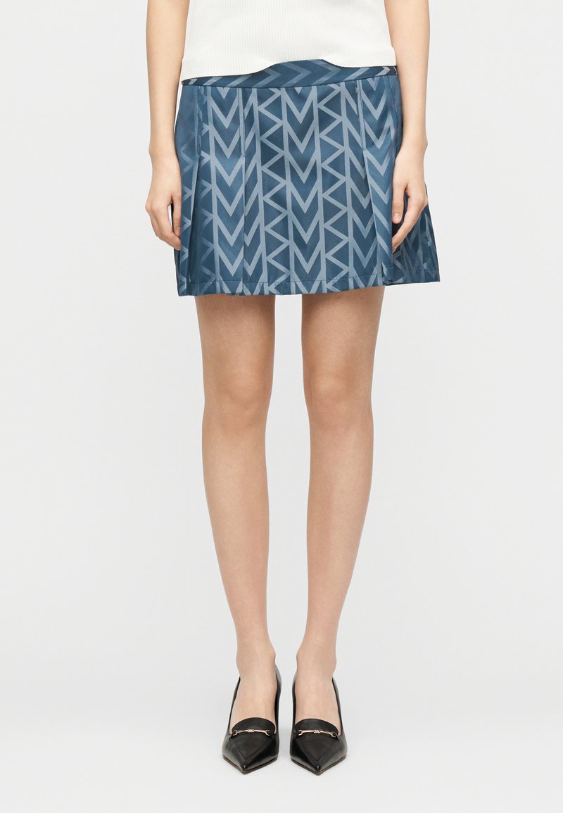 Γυναίκα που φοράει μια μπλε μίνι φούστα με σχέδια, συνδυασμένη με μαύρες μυτερές γόβες, στέκεται μπροστά σε ένα απλό ανοιχτό φόντο.