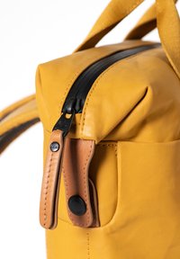 Gelber Stoffbeutel mit einem schwarzen Reißverschluss, einem beigen Lederakzent und einer abgerundeten Form. Enthält einen stabilen Stoffgriff und eine glatte Textur.