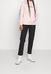 Felpa rosa con logo frontale, pantaloni neri e scarpe bianche alte con strisce nere. Lo zaino è di colore azzurro chiaro.