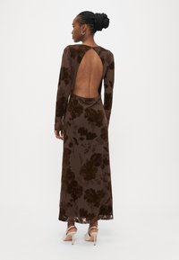 Μακρύ καφέ φόρεμα από βελούδο με λουλουδάτο σχέδιο, βαθύ άνοιγμα στην πλάτη, μακριά μανίκια και αέρινο σχέδιο. Λεπτή υφή και κομψή σιλουέτα.