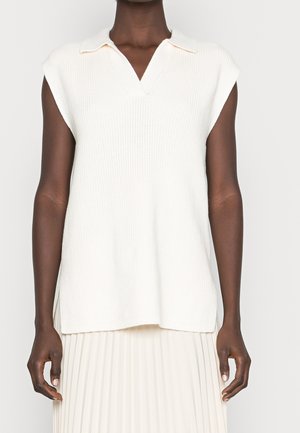 Osoba o ciemnej karnacji ubrana w bezrękawnik z białego dzianego materiału z kołnierzykiem oraz białą plisowaną spódnicę, stojąca na jednolitym tle.