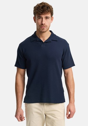 Hombre vistiendo una camisa polo de manga corta azul marino y pantalones beige claros, de pie frente a un fondo blanco liso.