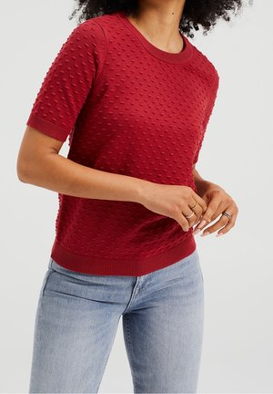 Kobieta w czerwonym swetrze o krótkich rękawach z teksturą i jasnoniebieskich dżinsach, stojąca z rękami złożonymi na wysokości talii.