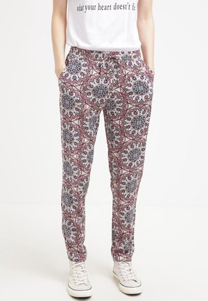 Kobieta w spodniach z troczkami we wzory z kołowymi i geometrycznymi motywami kwiatowymi, białe sportowe buty i biała koszula z częściowym tekstem.