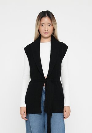 Zwarte gebreide vest met brede kraag en mouwloos ontwerp, met een riemomslag in de taille. Draag het over een wit langemouwen shirt en blauwe jeans.