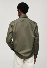 Olijfgroene, lange mouwen shirt met een achterpand, knoopsluiting kraag en een gladde stof. Draagt bij lichte broek.