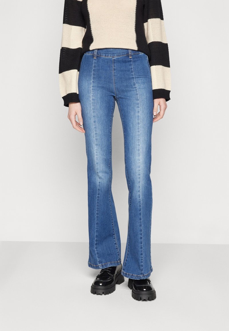 Femme portant un jean bootcut taille haute bleu, des chaussures noires épaisses et un pull beige et noir à rayures manches longues, debout devant un fond uni.