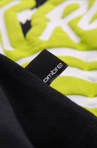 Tissu noir avec une petite étiquette tissée portant la mention « ombre » en blanc, superposée sur un fond textile graphique vert et blanc.