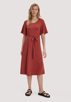 Frau trägt ein rostrotfarbenes Midikleid mit kurzen, ausgestellten Ärmeln und einem Taillengürtel, kombiniert mit schwarzen flachen Sandalen mit Riemchen, steht vor hellem Hintergrund.
