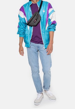 Giacca a vento azzurro chiaro con dettagli viola e bianchi, t-shirt viola, jeans azzurri chiari e sneakers beige, con una borsa a tracolla nera.