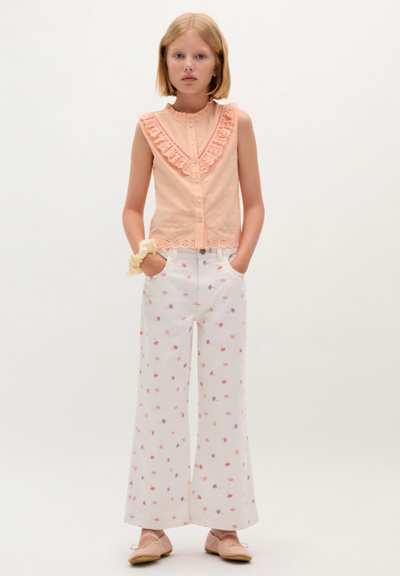 Pfirsichfarbenes Baumwollbluse mit Rüschensaum und Knopfverschluss, kombiniert mit weißen weiten Hosen mit floralem Muster in Orange und Lila.