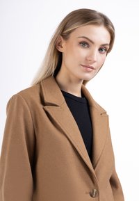 Blazer marrone in misto lana con rever a punta, chiusura a un bottone e texture morbida. Indossato sopra un top nero, evidenzia una vestibilità su misura.