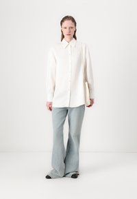 Camicia bianca con colletto, abbinata a jeans a campana blu chiaro e scarpe nere. Una piccola borsa bianca è indossata sulla spalla.