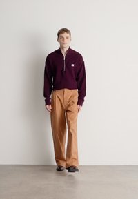 BURGUNDY, textúrált kötött pulóver negyed cipzáras gallérral, világos barna, bőszárú nadrággal és sötét cipővel párosítva. Semleges háttér.