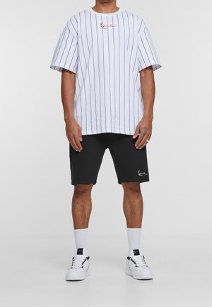 Weißes T-Shirt mit kurzen Ärmeln, schwarzen vertikalen Streifen und rotem Logo; kombiniert mit schwarzen Shorts und weißen Sneakers mit schwarzen Akzenten.