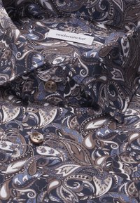 Chemise avec motif cachemire marron et bleu, boutons marron et une étiquette en tissu blanche lisant "seidensticker" à l'intérieur du col.