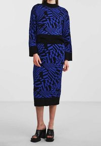 Sada pletených šatů s modrým a černým geometrickým vzorem. Dlouhé rukávy, kulatý výstřih a přiléhavý střih. Doplněno černými sandály na platformě.