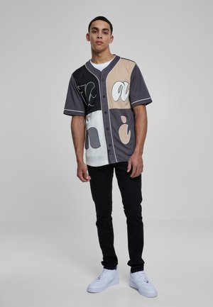 Camisa de botones de manga corta colorida con parches en beige, gris y blanco que presenta varios patrones. Llevada con pantalones negros y zapatos blancos.
