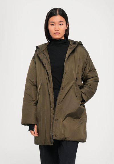 Frau in übergroßer, olivgrüner Kapuzenpuffjacke über einem schwarzen Rollkragenpullover, stehend vor hellem, einfarbigem Hintergrund.