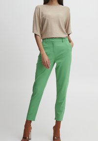 Una donna posa indossando un top beige e pantaloni verdi, con tacchi marroni. Si erge con sicurezza contro uno sfondo neutro.