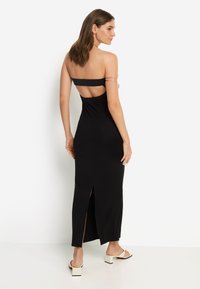 Μακρύ μαύρο φόρεμα ριμπ με μπαντό τοπ, ανοιχτή πλάτη και πλευρική σχισμή. Διαθέτει μίνιμαλ σχεδιασμό και απαλή υφή.