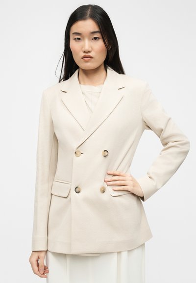 Γυναίκα με μακριά μαύρα μαλλιά, ντυμένη με κρεμ σακάκι διπλής σειράς με τέσσερα κουμπιά, στέκεται με το ένα χέρι στη μέση.