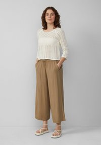 Lichtgewicht, crème kleurige gebreide trui met een openweefpatroon, gecombineerd met losse, tan kleurige broeken met een elastische tailleband en zakken. Witte sandalen.