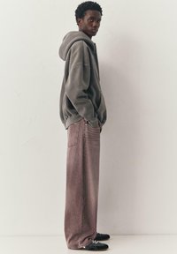 Jeune personne portant un sweat à capuche gris oversize, un jean rose délavé à larges jambes et des baskets noires, debout de profil contre un mur blanc uni.