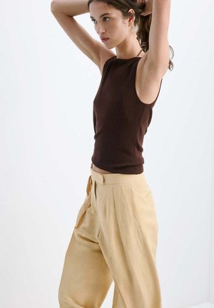 Femme ajustant ses cheveux, portant un haut marron foncé sans manches et un pantalon large beige taille haute sur un fond uni.