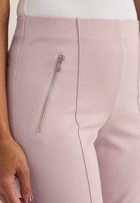 Nahaufnahme einer Person, die hoch taillierte rosa Hosen mit einer vertikalen silbernen Reißverschlusstasche an der Seite und ein weißes geripptes Oberteil trägt.