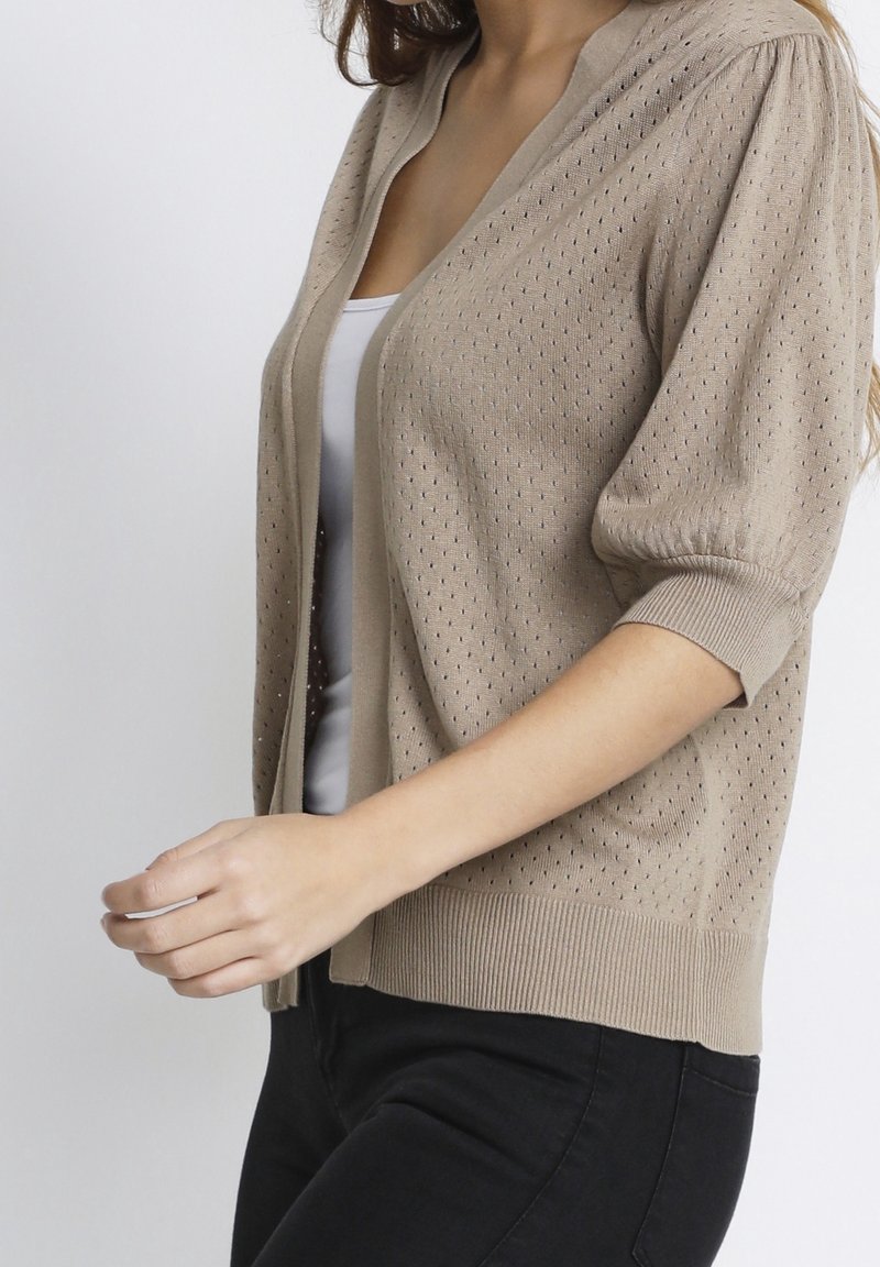 Beige stickad cardigan med ett mönster, korta ärmar och ribbade muddar. Har en öppen front och en avslappnad passform.