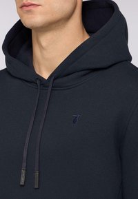 Mörkblå hoodie i en bomullsblandning med en känguruficka, snörning och en diskret broderad logga på bröstet.