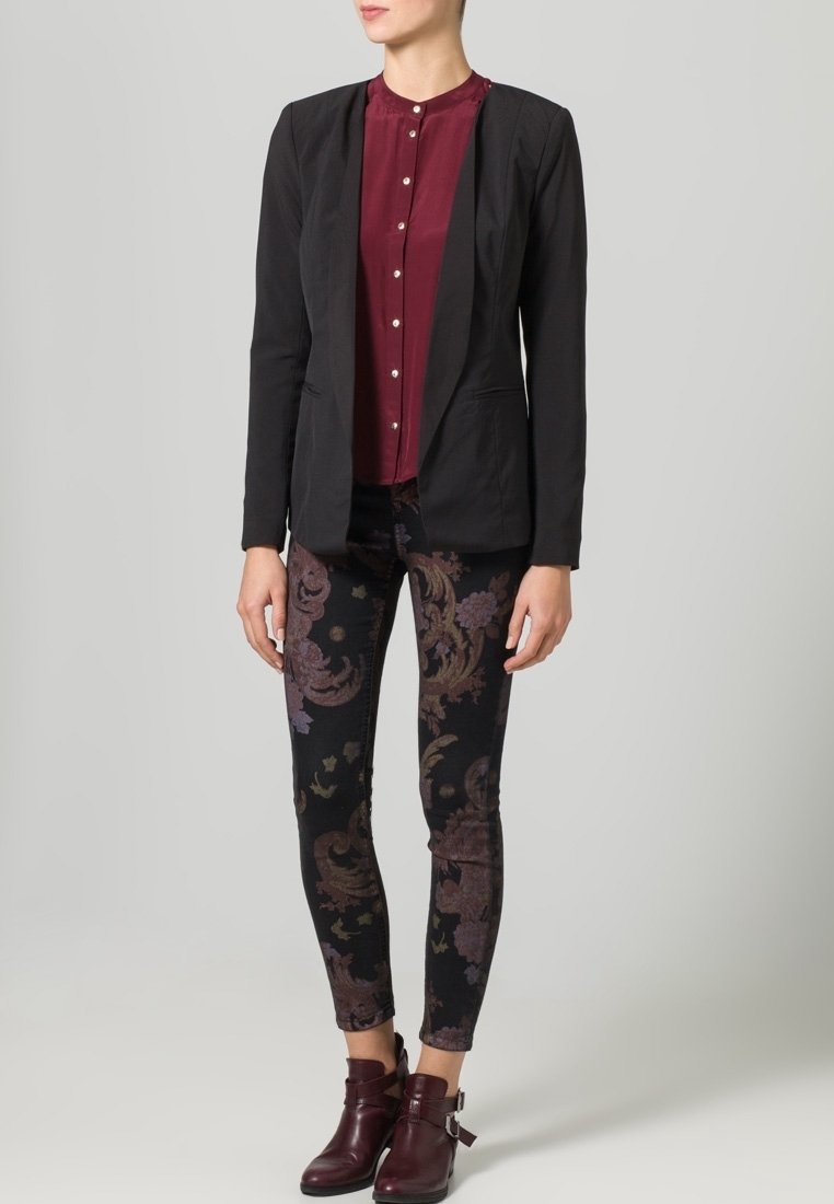 Femme portant un blazer noir, un chemisier bordeaux boutonné, un pantalon skinny sombre à motifs et des bottines bordeaux.