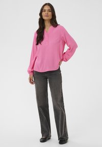 Femme aux longs cheveux noirs portant un blouse à manches longues rose vif, un jean gris foncé et des chaussures noires, se tenant avec une main sur la hanche.