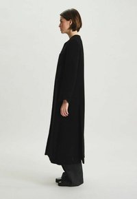 Manteau noir long avec une silhouette droite, en tissu texturé, doté de longues manches et sans fermetures visibles.