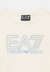 Kreminės spalvos medvilniniai marškinėliai su apvalia iškirpte, priekyje su šviesiai mėlynu "EA7 EMPORIO ARMANI" logotipo spaudu.