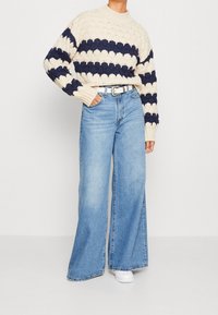 Henkilö, joka on pukeutunut vaalean kermanväriseen ja laivastonsiniseen raidoitettuun neulepaitaan, korkeavyötäröisiin vaaleansinisiin leveisiin farkkuihin, valkoiseen vyöhön ja valkoisiin tennareihin, seisoo rentona.