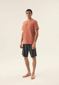 Czerwono-pasiasty bawełniany t-shirt z dekoltem w serek, połączony z szarymi szortami. Koszulka na środku ma logo i jest noszona na boso.